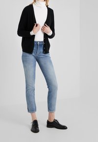 Donna che indossa un cardigan nero con bottoni sopra un dolcevita bianco, jeans azzurri chiari e scarpe nere con lacci, in piedi contro uno sfondo bianco uniforme.