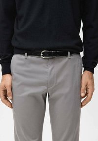 Maglione nero a maglia indossato con pantaloni grigi. Una cintura nera in pelle con fibbia argento tiene i pantaloni, mostrando una texture liscia.