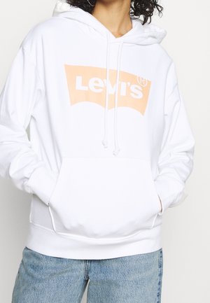 Osoba nosząca biały kaptur Levi's z logo brzoskwini, ręce w przedniej kieszeni, połączony z jasnoniebieskimi dżinsami.