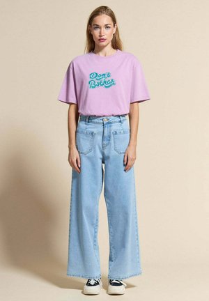 Jeune femme debout, face à l'avant, portant un t-shirt lavande avec l'inscription « Ne Pas Déranger », un jean large bleu clair et des baskets noires et blanches.