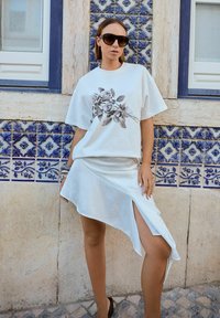 Tricou alb cu un print floral negru, asortat cu o fustă albă asimetrică din satin și ochelari de soare negri. Fundal cu faianță din ceramică.