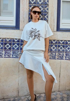 Tricou alb cu un print floral negru, asortat cu o fustă albă asimetrică din satin și ochelari de soare negri. Fundal cu faianță din ceramică.