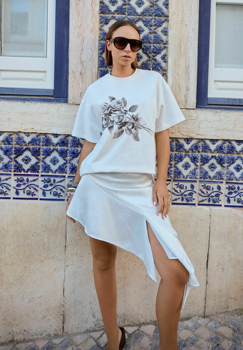 Tricou alb cu un print floral negru, asortat cu o fustă albă asimetrică din satin și ochelari de soare negri. Fundal cu faianță din ceramică.