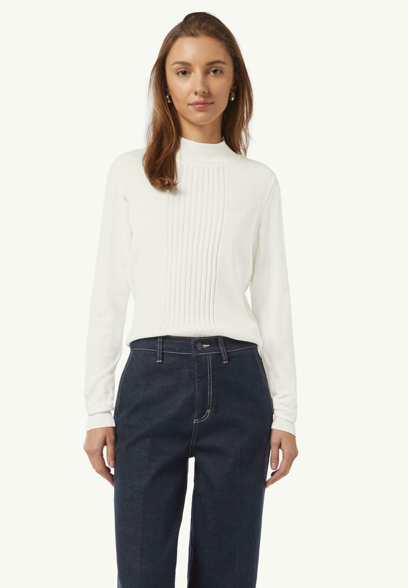 White ribbed sweater with a high neckline and vertical texture details, paired with dark blue high-waisted denim pants featuring contrasting stitching.