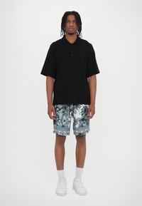 Polo shirt nero a maniche corte, in misto cotone, abbinato a pantaloni da nuoto bianchi e blu a fantasia. Sneakers bianche e calzini grigi alla caviglia.