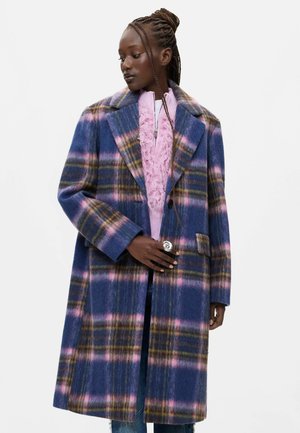 Donna con capelli intrecciati che indossa un cappotto a quadri blu, rosa e marrone sopra un maglione rosa con zip strutturato.