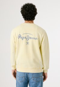 Geel katoenen sweatshirt met lange mouwen, ribgebreide manchetten en een groot blauw logo op de rug. Draagt een lichtblauwe denim jeans.