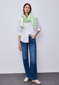 Femme debout contre un mur, portant une chemise blanche, une écharpe verte et blanche, un jean bleu et des baskets beiges, une main dans la poche.