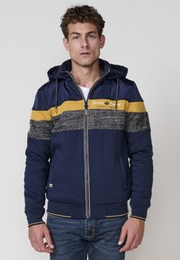 Sudadera con cremallera en color navy, diseño de rayas amarillas y grises, textura suave y capucha ajustable. Cuenta con bolsillos laterales y puños de canalé.