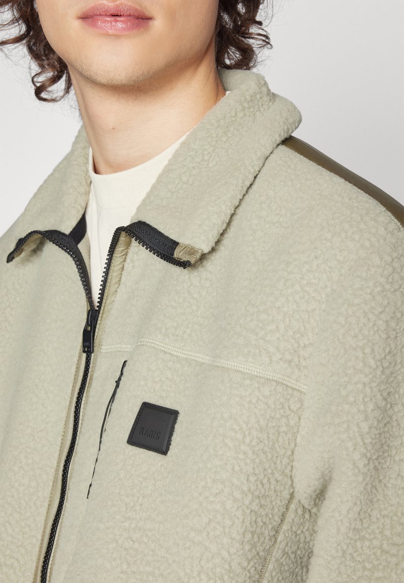 Ljusgrön fleecejacka med dragkedja fram, krage, svarta detaljer och en liten svart etikett. Texturerad tyg och kontrasterande axeldetaljer.