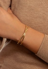Goldbangle-Armband mit doppelter Twist-Design, glatter Textur und glänzender Oberfläche, getragen an einer Hand gegen einen hellbraunen Strickstoff.