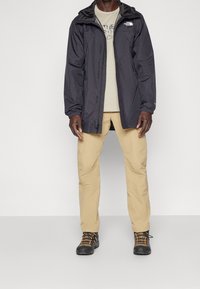 Svart vattentät jacka med huva över en beige t-shirt, tillsammans med ljusbeiga byxor och svarta vandringsskor med orange detaljer.