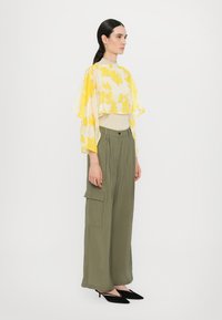 Gul bluse med blomsterprint og flæseærmer, parret med olivengrønne bukser med vide ben. Modellen har sorte spidse hæle på.