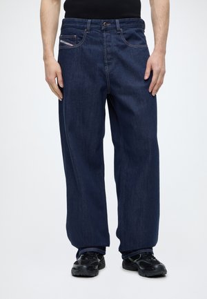 Homme portant un jean ample bleu foncé avec des baskets noires et une chemise noire, debout devant un fond blanc uni.