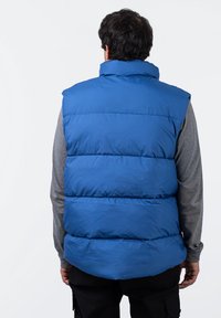 Chaleco acolchado aislante azul con cuello alto, que presenta paneles cosidos. El fondo es blanco liso y el modelo lleva una camiseta de manga larga gris.