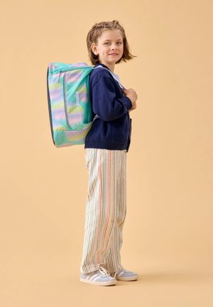 Backpack with a multicolored bubble pattern, made of lightweight material. Model wears a navy cardigan and striped pants, with gray sneakers.