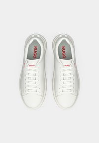White leather sneakers with round toes, six pairs of eyelets, and red "HUGO" branding on the sides and tongue. Textured rubber sole.