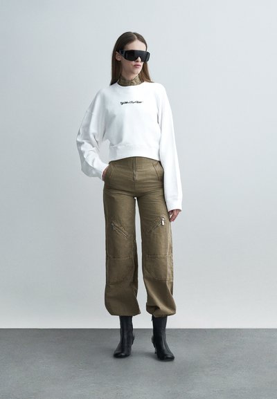 Femme debout portant un sweat-shirt blanc oversize, un pantalon cargo vert olive, des bottes noires à talons et de grandes lunettes de soleil noires sur un fond uni.