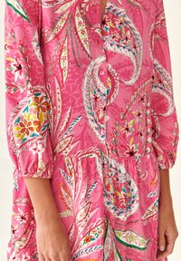 Abito rosa brillante con motivi floreali e paisley colorati, con bottoni sul davanti e maniche a sbuffo a tre quarti, indossato da una persona.