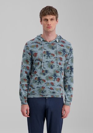 Junger Mann trägt einen hellblauen Kapuzenpullover mit Blumendesign und Kordeln sowie eine marineblaue Hose und steht vor einem einfarbigen grauen Hintergrund.
