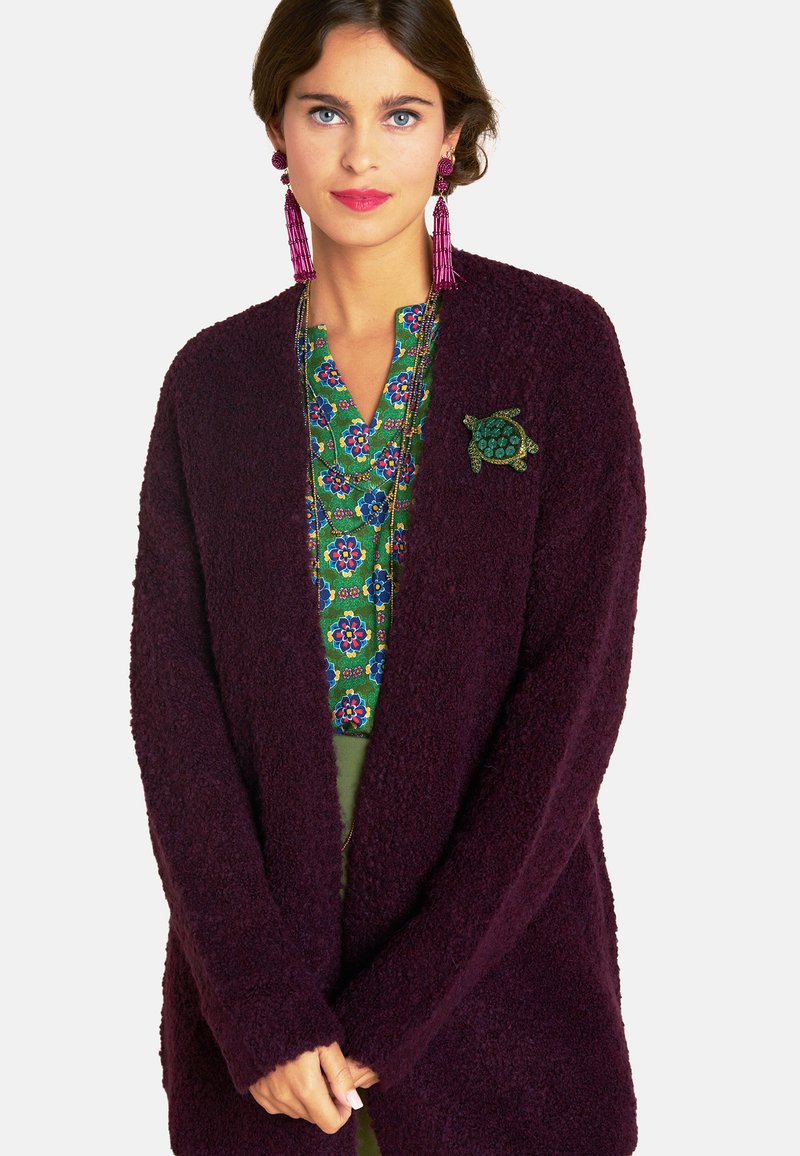 Eine Frau trägt eine lila strukturierte Strickjacke mit einer Schildkrötenbrosche, eine grüne Bluse mit Blumenmuster, eine lange Perlenkette und rosa Quastenohrringe.