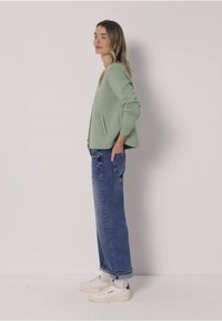 Suéter texturizado verde claro de manga larga, combinado con jeans azules holgados y zapatillas blancas con detalles grises. Vista lateral mostrada.