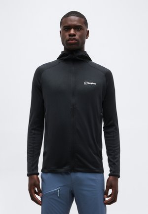 Schwarze Zip-up-Jacke mit Kapuze, getragen von einem stehenden Mann, kombiniert mit hellblauen Hosen mit einem Reißverschlusstaschen am Oberschenkel.