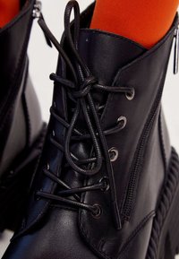 Black leather ankle boot with a round toe, lace-up front, zip closure, and textured rubber sole. Features metallic eyelets and flat laces.