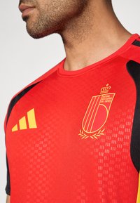 Raudona sportinė marškinė, kuriuose yra juodų akcentų, tekstūruotas audinys, geltona Belgijos futbolo asociacijos logotipas ir trumpomis rankovėmis.