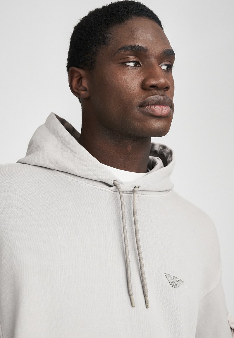 Tânărul bărbat de culoare purtând un hanorac gri deschis cu șnururi, uitându-se în lateral, pe un fundal alb simplu.