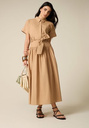 Frau trägt ein beiges kurzärmeliges Hemdkleid mit gebundener Taille, schwarze Riemchensandalen, braune Armbänder, hält drei blasse Handtaschen.