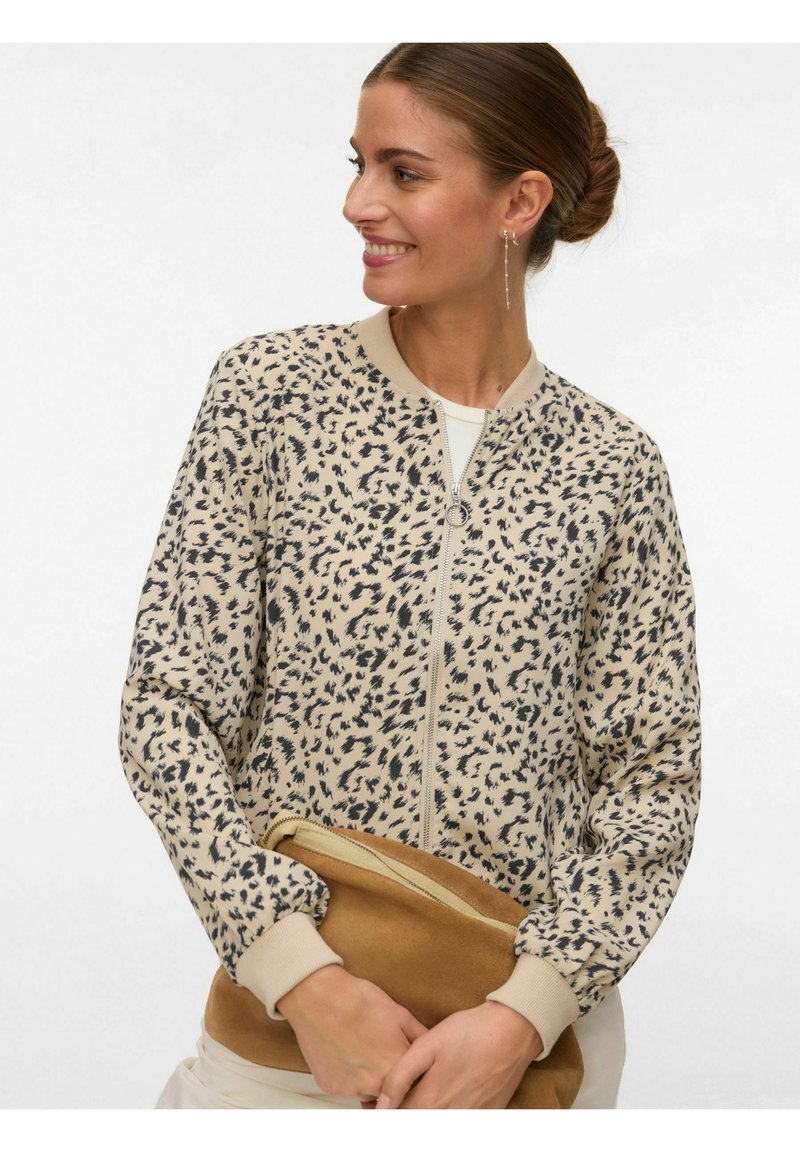 Femme souriante, portant une veste zippée en imprimé léopard beige et noir, tenant une grande pochette en daim marron, cheveux attachés en chignon.