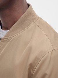Gros plan d'une personne portant une veste bomber beige avec un col côtelé sur une chemise gris clair.