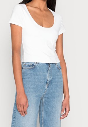 Kvinde iført en tætsiddende hvid top med rund hals og korte ærmer, kombineret med lyseblå, højtaljede denimjeans.