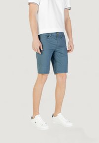 Bermudas azules para hombre fabricadas en un material ligero, con cremallera en la bragueta, bolsillos frontales y costuras planas. Combinadas con zapatillas blancas.