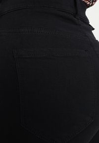 Acercamiento de un bolsillo trasero de unos vaqueros negros con costuras visibles y parte de una tela floral encima de la cinturilla.