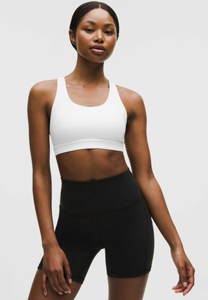 Zadbana kobieta o ciemnej karnacji, ubrana w biały sportowy biustonosz i czarne, wysokowciskane szorty rowerowe, stojąca na jasnym, jednolitym tle.