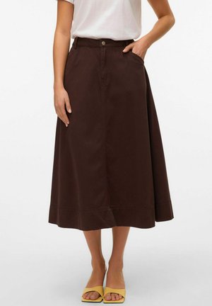 Donna che indossa una gonna midi A-line marrone scuro con tasche, una maglietta bianca infilata dentro e sandali gialli con tacco a blocco aperti.