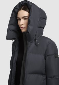 Chaqueta de plumas en gris oscuro con una capucha grande y completa. Cuenta con un cierre de cremallera en la parte delantera, tejido texturizado y un acento de logo circular en el brazo izquierdo.