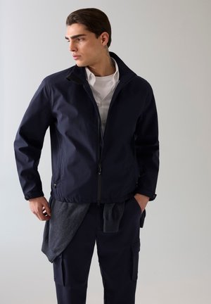 Homme portant une veste zippée bleu marine sur une chemise blanche, avec un pull gris noué autour de la taille, regardant vers la gauche sur un fond uni.
