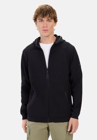 Schwarze Zip-Hoodie mit einer Kordelzugkapuze, gerippter Textur, zwei vorderen Taschen und kontrastierenden Reißverschlusdetails, getragen über einem weißen Shirt.