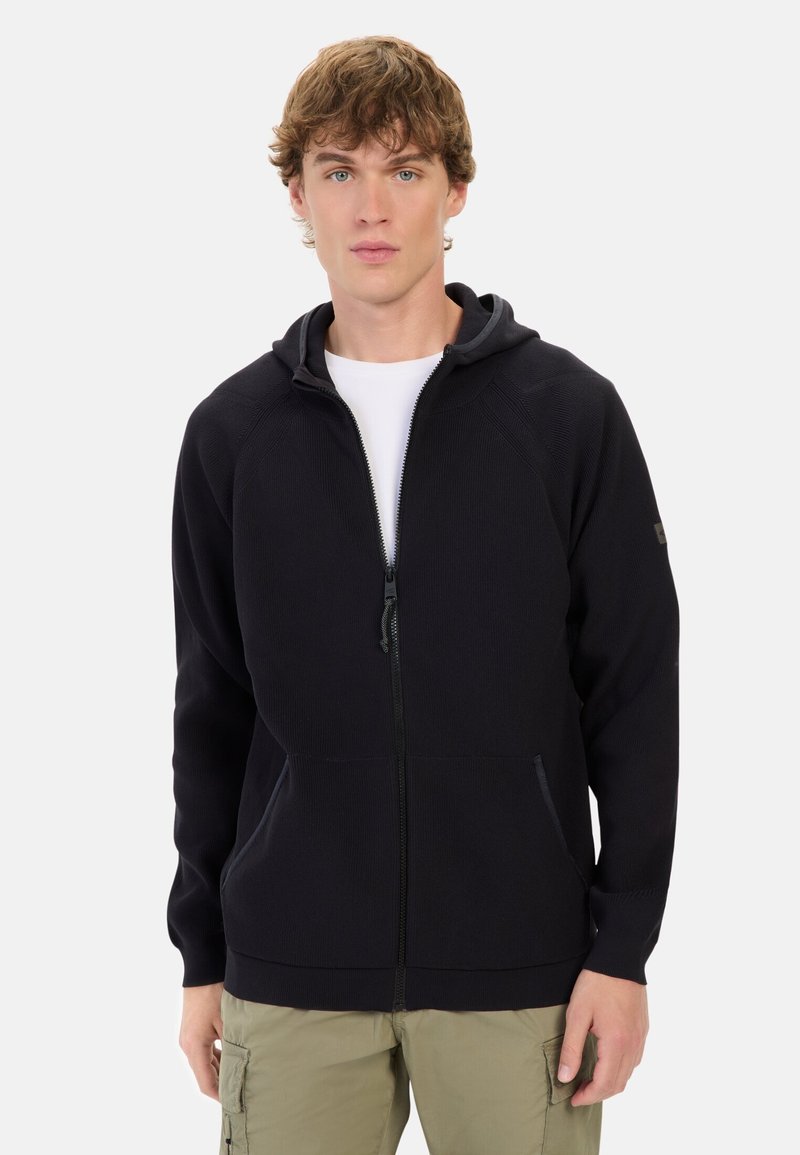 Schwarze Zip-Hoodie mit einer Kordelzugkapuze, gerippter Textur, zwei vorderen Taschen und kontrastierenden Reißverschlusdetails, getragen über einem weißen Shirt.