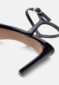 Μαύρο σανδάλι με τακούνι από πατεντέ δέρμα, λεπτό λουράκι στον αστράγαλο και χρυσή αγκράφα, σε γωνία σε λευκό φόντο.