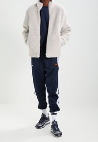 Fleece zip-jacka i mjuk krämfärg, ihopparad med marinblå träningsbyxor med vita sidostråk och en logotyp. Svarta sneakers med silverdetaljer.