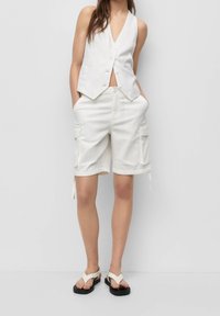 Gilet bianco in lino con scollatura a V e bottoni, abbinato a pantaloni cargo bianchi con tasche laterali. Indossato con sandali infradito bianchi.