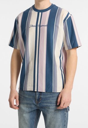 Mężczyzna w krótkim rękawie koszulki w pionowe paski w kolorach niebieskim, kremowym i różowym z napisem "Guess Originals", ubrany w niebieskie dżinsy.