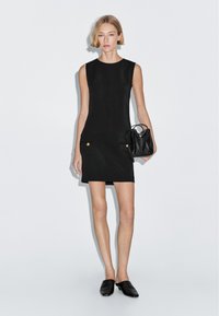 Schwarzes ärmelloses Kleid mit taillierter Form, ausgestattet mit zwei vorderen Taschen mit goldenen Knöpfen. Kombiniert mit einer kleinen schwarzen Handtasche und flachen Schuhen.