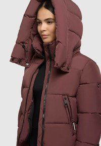 Hnědá péřová bunda s velkou kapucí, obsahující dvoucestný zip, kapsu na zip a prošívaný design pro izolaci a teplo.