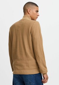 Beige strukturierter Pullover mit hohem Kragen, gerippten Bündchen und Saum, tailliertem Design und einem gewebten Muster. Die Ansicht ist von hinten.
