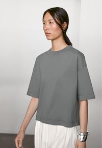 Grå oversized T-shirt lavet af bomuld, med korte ærmer, rund halsudskæring og lige kant. Modellen bærer sølvarmbånd på håndleddet.